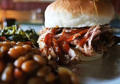 A close-up of a pulled pork sandwich with barbecue sauce, served with baked beans and greens on a plate.