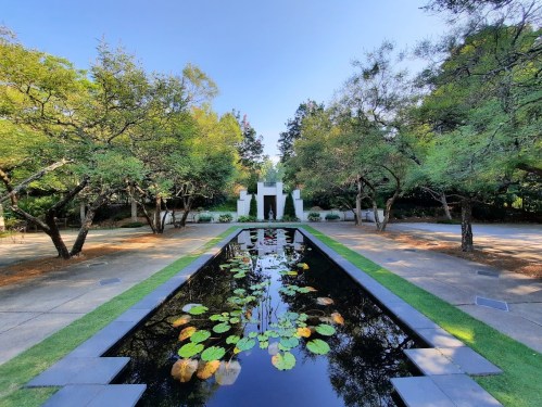 A serene garden with a reflecting pool, surrounded by trees and white sculptures under a clear blue sky.