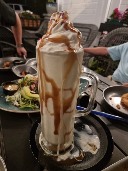 A tall glass of milkshake topped with whipped cream and caramel drizzle, served on a plate with food in the background.