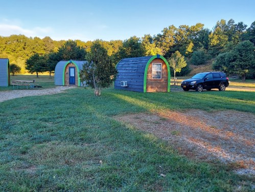 Two small, colorful cabins in a grassy area, with a black SUV parked nearby and trees in the background.