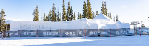 A snowy landscape featuring a large snow structure with "Keystone" signage, surrounded by a fence and pine trees.
