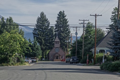 A quaint church surrounded by trees on a quiet street, with power lines and distant mountains in the background.
