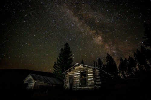 A rustic cabin under a starry night sky, with the Milky Way visible above and trees in the background.