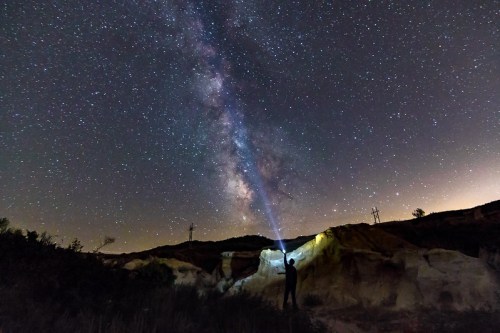 A person stands in a dark landscape, pointing a flashlight at the starry sky, showcasing the Milky Way.
