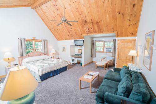 Cozy room with a high wooden ceiling, king bed, seating area, and a small TV, featuring warm lighting and carpeted floor.