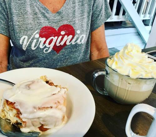 A person in a "Virginia" shirt enjoys a cinnamon roll with icing and a coffee topped with whipped cream.
