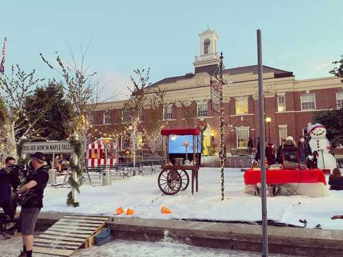 A festive outdoor scene with snow, decorations, a snowman, and a building in the background, set for a winter event.