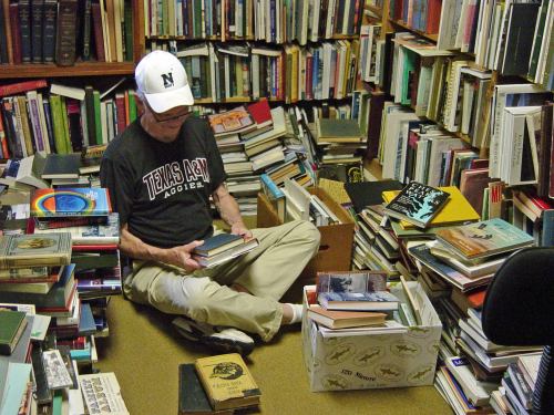 A person sits on the floor surrounded by stacks of books, examining one in a cluttered room filled with shelves.
