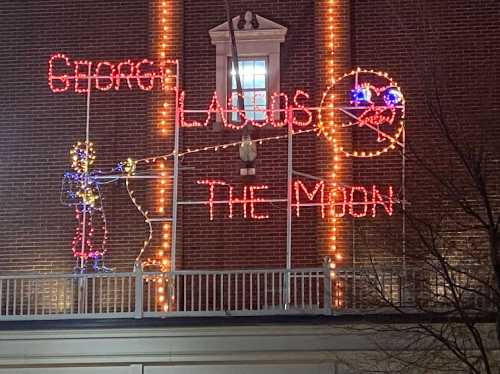 Colorful holiday lights display featuring the text "George" and "The Moon," with a figure casting a fishing line.