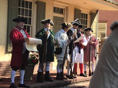 Five individuals in historical costumes stand outside a building, holding documents and speaking to an audience.