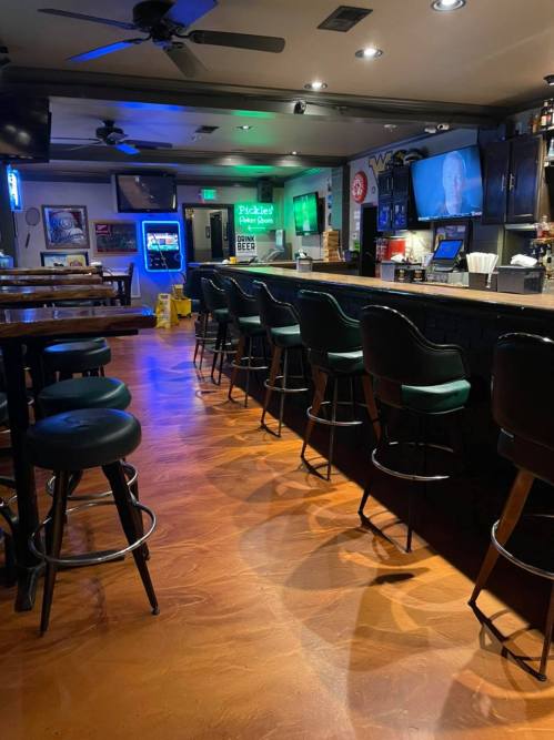 Interior of a bar with empty stools, a long counter, and dim lighting, featuring a jukebox and a TV in the background.