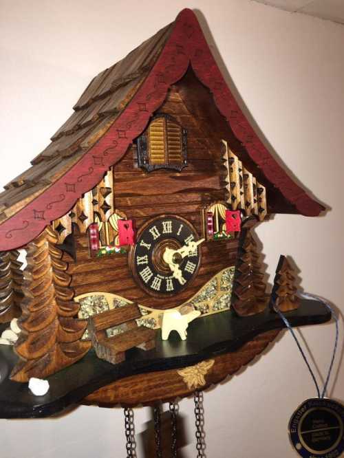 A decorative wooden cuckoo clock with a chalet design, featuring trees and a clock face with Roman numerals.