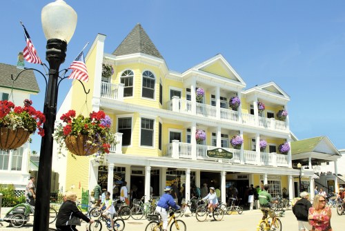 A sunny street scene featuring a yellow building with balconies, flower baskets, and people biking and walking.