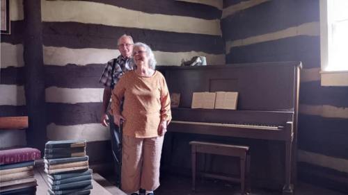 An elderly couple stands in a rustic room with a piano and stacked books, smiling and enjoying each other's company.