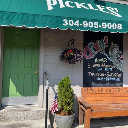 A storefront with a green awning reading "Pickles!" and an "Open" sign, featuring outdoor seating and a flag.