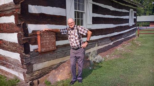 An elderly man in a plaid shirt stands beside a log cabin with a wooden sign, smiling at the camera.
