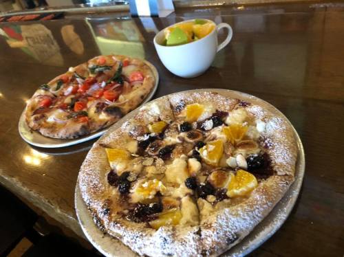 Two pizzas on metal plates: one with tomatoes and greens, the other with fruit and powdered sugar, alongside a cup of fruit.