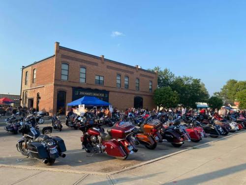 A gathering of motorcycles parked outside a brick building, with people mingling and a blue tent in the background.