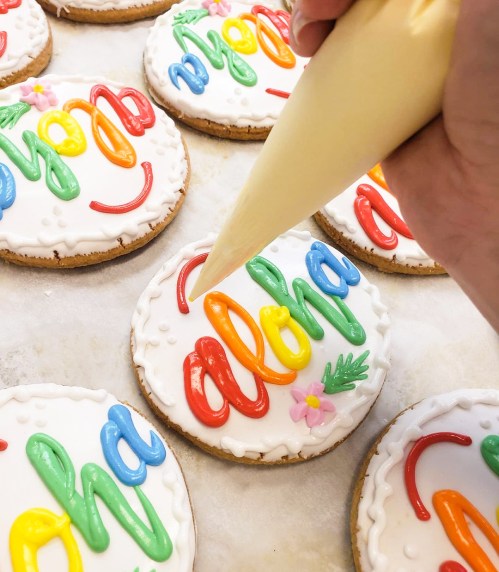 A hand decorates cookies with colorful icing, spelling "aloha" on each cookie.