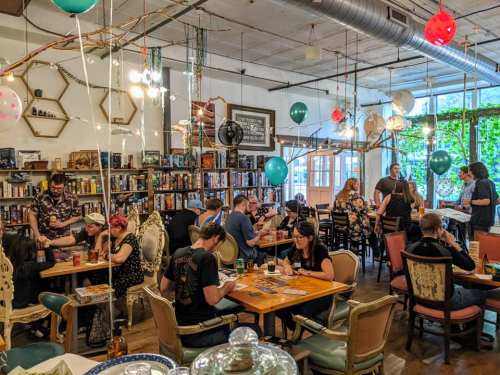 A lively board game café with people playing games, colorful balloons, and shelves filled with board games.