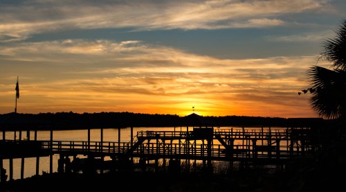 Sunset over a calm lake, with silhouettes of a dock and palm trees against a colorful sky.