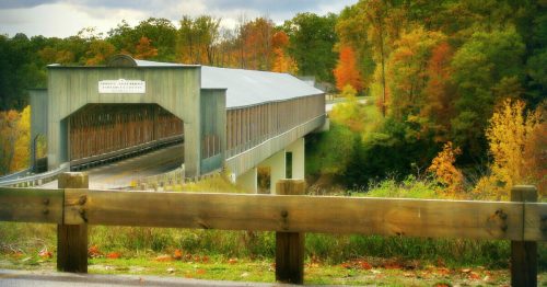 A scenic view of a covered bridge surrounded by colorful autumn foliage and a wooden fence in the foreground.