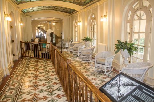 A well-lit hallway with a patterned tile floor, featuring wicker chairs and potted plants, framed by arched windows.