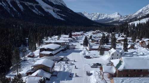 A snowy mountain town with wooden buildings, surrounded by tall trees and mountains under a clear blue sky.