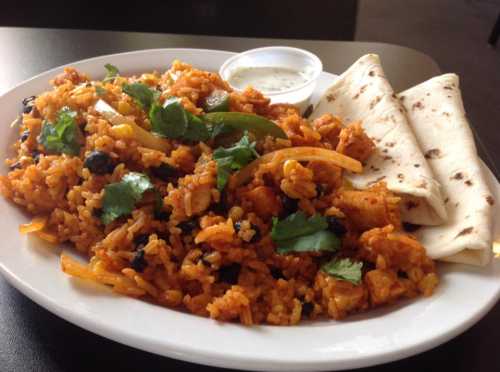 A plate of flavorful rice with vegetables and chicken, served with two flatbreads and a small bowl of sauce.