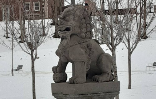 A stone lion statue sits on a pedestal in a snowy park, surrounded by bare trees and benches.