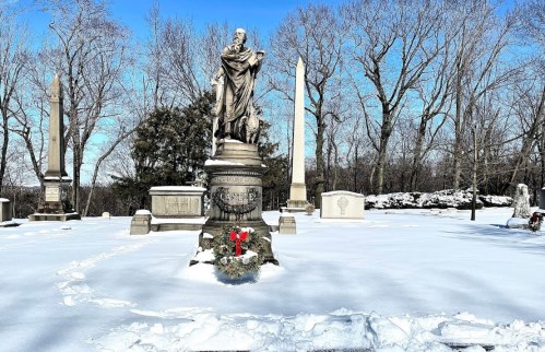 A snowy cemetery scene featuring a statue, obelisks, and a wreath on a grave, surrounded by bare trees.