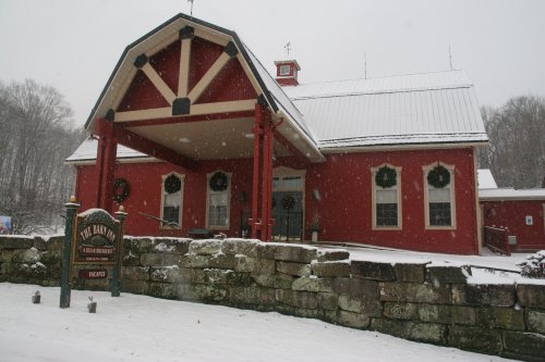 A red barn-style building covered in snow, with a stone wall and holiday decorations visible. Snowflakes are falling.