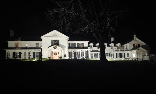 A large, illuminated white house at night, featuring a festive wreath and surrounded by dark trees.