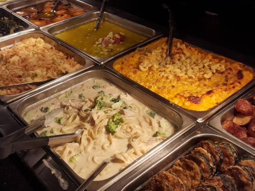 A buffet display featuring various dishes, including pasta, macaroni and cheese, rice, and vegetables in trays.