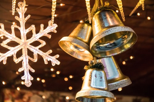 Decorative golden bells and a clear snowflake hang against a backdrop of warm, twinkling lights.