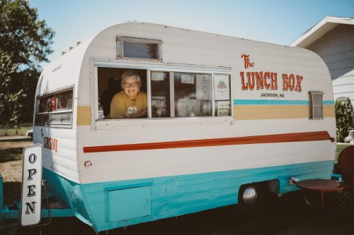 A cheerful vendor smiles from the window of a vintage food trailer named "The Lunch Box."