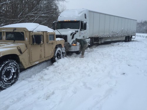 A military Humvee assists a stuck semi-truck in deep snow during winter weather.