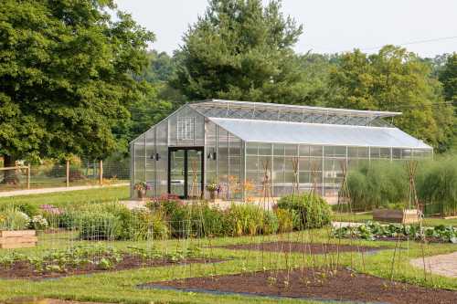 A greenhouse surrounded by lush greenery and garden beds, set against a backdrop of trees and a clear sky.