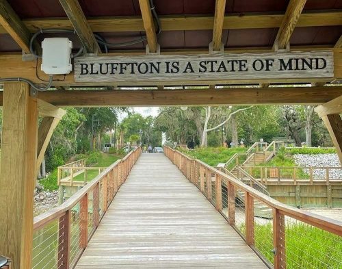 A wooden bridge leads to a path with a sign reading "Bluffton is a state of mind" above. Lush greenery surrounds the area.