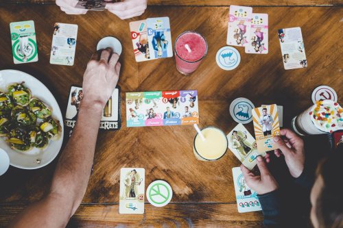 A tabletop scene with hands playing card games, surrounded by drinks and snacks. Colorful cards are spread out.