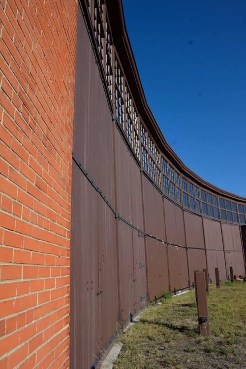 Curved wooden wall with large windows, adjacent to a brick structure, under a clear blue sky.