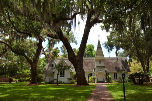 A charming church surrounded by lush greenery and large trees draped with Spanish moss under a clear blue sky.