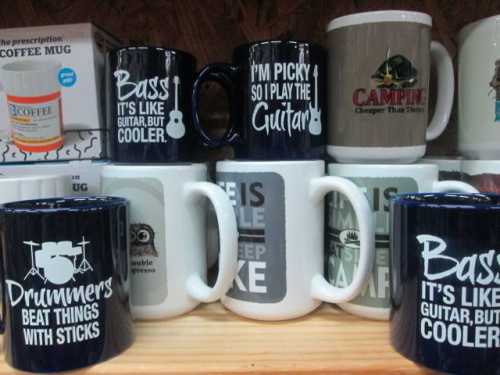 A collection of mugs with humorous music-themed quotes about guitars and drumming, displayed on a shelf.