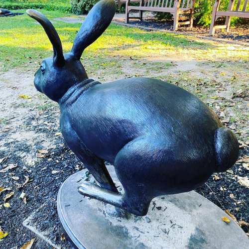A large, black rabbit sculpture sits on a circular base in a park, surrounded by grass and benches.