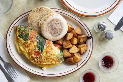 A plate with a vegetable omelet, roasted potatoes, and an English muffin, accompanied by small dishes of jam.