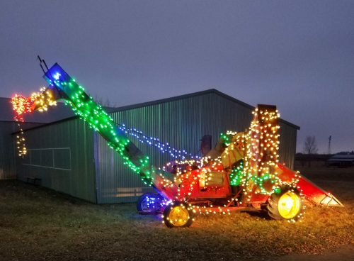 A brightly lit farm machine decorated with colorful Christmas lights, set against a gray sky and a metal shed.