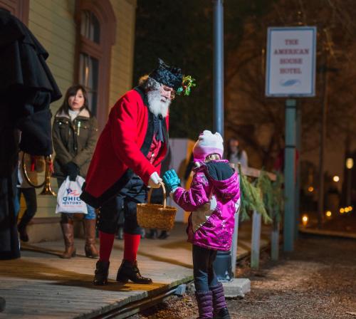 A man in a red coat and hat hands a basket to a young girl in winter attire outside a hotel during a festive event.
