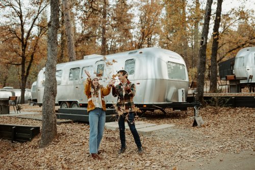 Two people joyfully toss autumn leaves in front of a silver Airstream trailer, surrounded by trees and fallen leaves.
