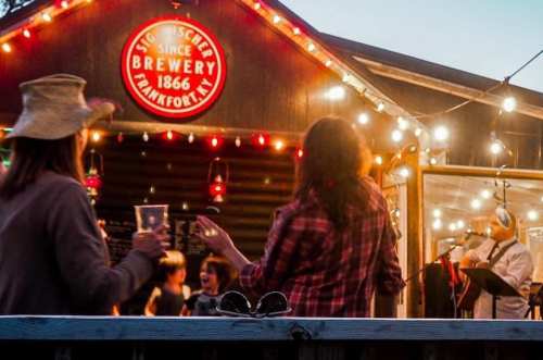 People enjoying a lively outdoor event at a brewery, with string lights and a musician performing.