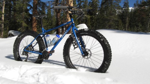 A blue fat bike stands in deep snow, surrounded by trees and a clear blue sky.